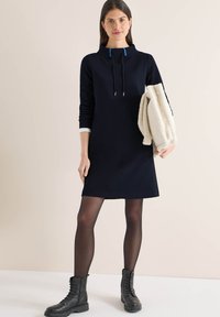 Robe droite bleu marine avec un col montant, des cordons de serrage ajustables et des poignets blancs contrastants, portée avec un manteau beige et duveteux.