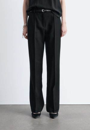 Czarne, dopasowane spodnie z wysokim stanem i przednią plisą, noszone z cienkim czarnym paskiem, czarnymi butami z szpiczastym noskiem oraz srebrnymi bransoletkami i pierścionkami.