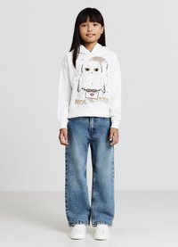 Un sweat à capuche blanc avec un graphique de hibou, "Hogwarts" en texte doré, associé à un jean en denim large bleu clair avec des studs décoratifs. Des baskets blanches.