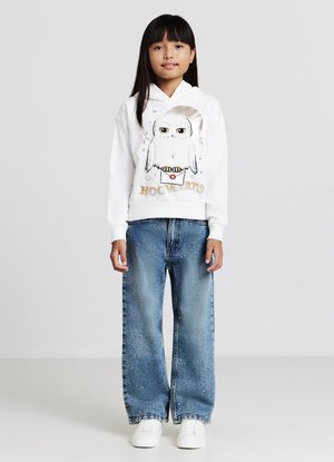 Un sweat à capuche blanc avec un graphique de hibou, "Hogwarts" en texte doré, associé à un jean en denim large bleu clair avec des studs décoratifs. Des baskets blanches.
