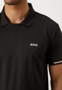 Fekete póló, textúrált felülettel, három gombos nyakkal és fehér "BOSS" logóval a mellrészen. A gallér csíkos elemeket tartalmaz.