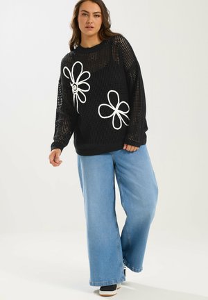 Kvinde iført sort åbenstrikket trøje med hvide blomstermotiver og løstsiddende lyseblå jeans, stående mod en ensartet lys baggrund.