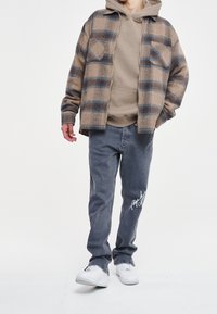 Brązowa i niebieska koszula w kratę flanelową na beżowym hoodie, w połączeniu z jasnoszarymi dżinsami z białym haftem i białymi sneakersami.