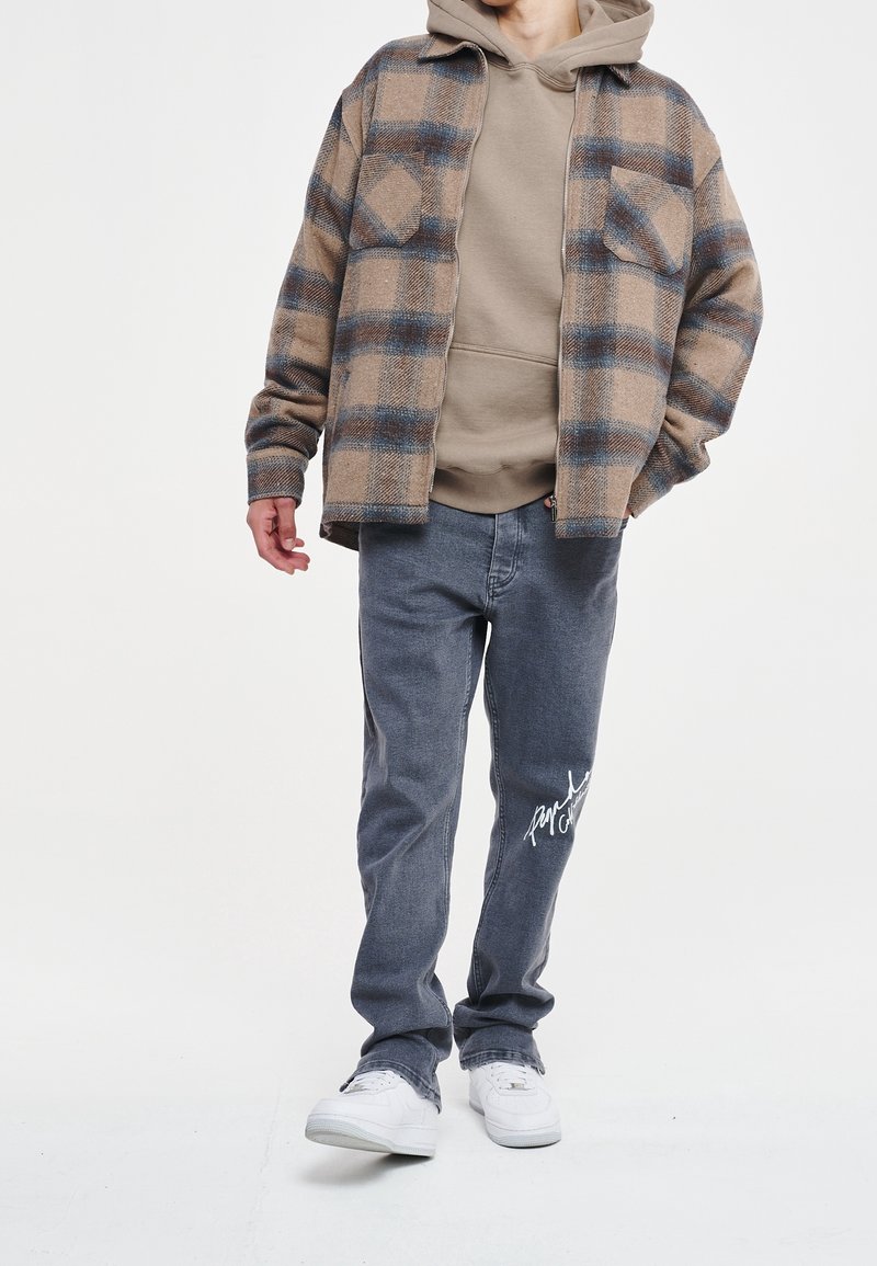Brązowa i niebieska koszula w kratę flanelową na beżowym hoodie, w połączeniu z jasnoszarymi dżinsami z białym haftem i białymi sneakersami.