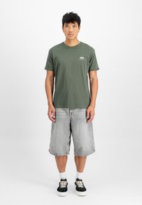 Olivengrønn T-skjorte med korte ermer og rund hals, med lite grafikk på venstre bryst, parret med lysegrå denimshorts og svarte joggesko.