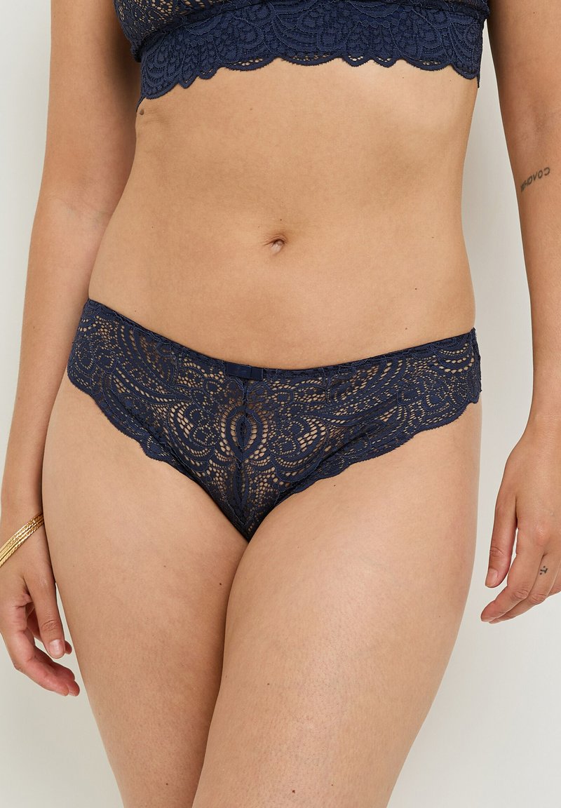 Donna che indossa un reggiseno e delle mutande in pizzo blu scuro abbinati, in piedi davanti a uno sfondo neutro con tatuaggi visibili sulle braccia e un braccialetto.