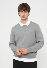 Γκρι φούτερ με στρογγυλή λαιμόκοψη, ριμπ μανσέτες και τελείωμα. Φοράει λευκό πουκάμισο με γιακά από μέσα. Απλή επωνυμία στο στήθος.