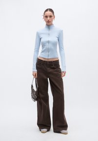 Ανοιχτό μπλε, μακρυμάνικο τοπ με φερμουάρ, σετ με φαρδιά, σκούρα καφέ παντελόνια. Κρατά μικρή μαύρη τσάντα με αλυσίδα.