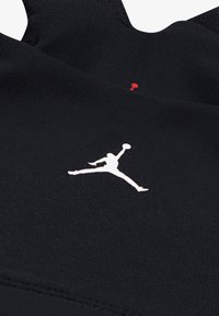 Melnā sportiskā apģērba gabals ar baltu silueta logo, ribotu tekstūru un sarkanu akcentu detaļu pie pleca.