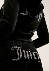 Fekete bársonyból készült melegítőszett, amelyen a hátán strasszokkal díszített "Juicy" logó található, kapucnival és slim-fit fazonnal.