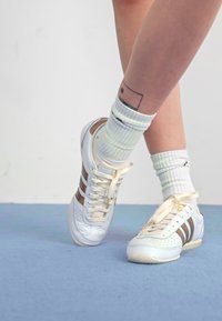 Adidași albi cu dungi maro, având un design perforat, șireturi bej și o talpă texturată. Detaliați cu accente aurii.