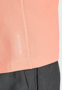 Camisa transpirable de color rosa claro con un diseño de malla texturizada, que presenta un dobladillo cosido y la palabra "AEROREADY" en azul claro cerca de la parte inferior.