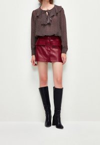 Blusa negra con lunares y detalles de volantes, combinada con una mini falda de cuero burdeos que cuenta con bolsillos frontales y botones. Botas de tacón altas negras.