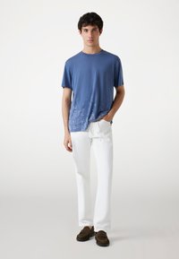 Camiseta azul de manga corta con un diseño de paisley en el dobladillo, combinada con pantalones blancos de pierna recta y zapatos marrones sin cordones.