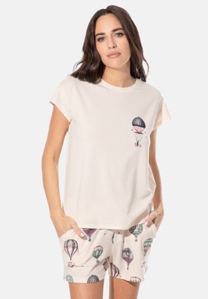 Woman wearing cream-colored top with hot air balloon graphic and matching shorts printed with various hot air balloons, hands in pockets.