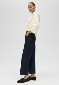Vrouw draagt een crème-kleurige capuchontrui, donkere wijde jeans en donkere mocassins, staand met handen in zakken.