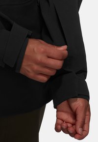 Svart jacka med en närbild på en hand som justerar en manschett. Materialet är slätt, har en dragkedjedetalj och en avslappnad passform.