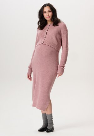 Geribbelde gebreide, roze cardigan met knopen, gecombineerd met een roze aansluitende jurk. Gekreukt grijze sokken en zwarte schoenen maken de look compleet.