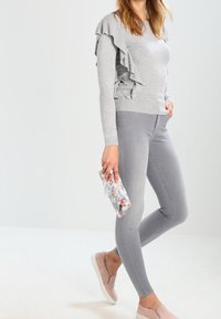 Felpa grigia con dettagli arricciati sul lato, abbinata a jeans aderenti grigio chiaro e scarpe slip-on color blush. Tienendo in mano una pochette con motivo floreale.