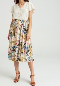 Lichte, beige blouse met korte, gerimpelde mouwen, gecombineerd met een bloemenrok tot de knie met blauwe, roze en gele patronen. Bruine, hoge sandalen.