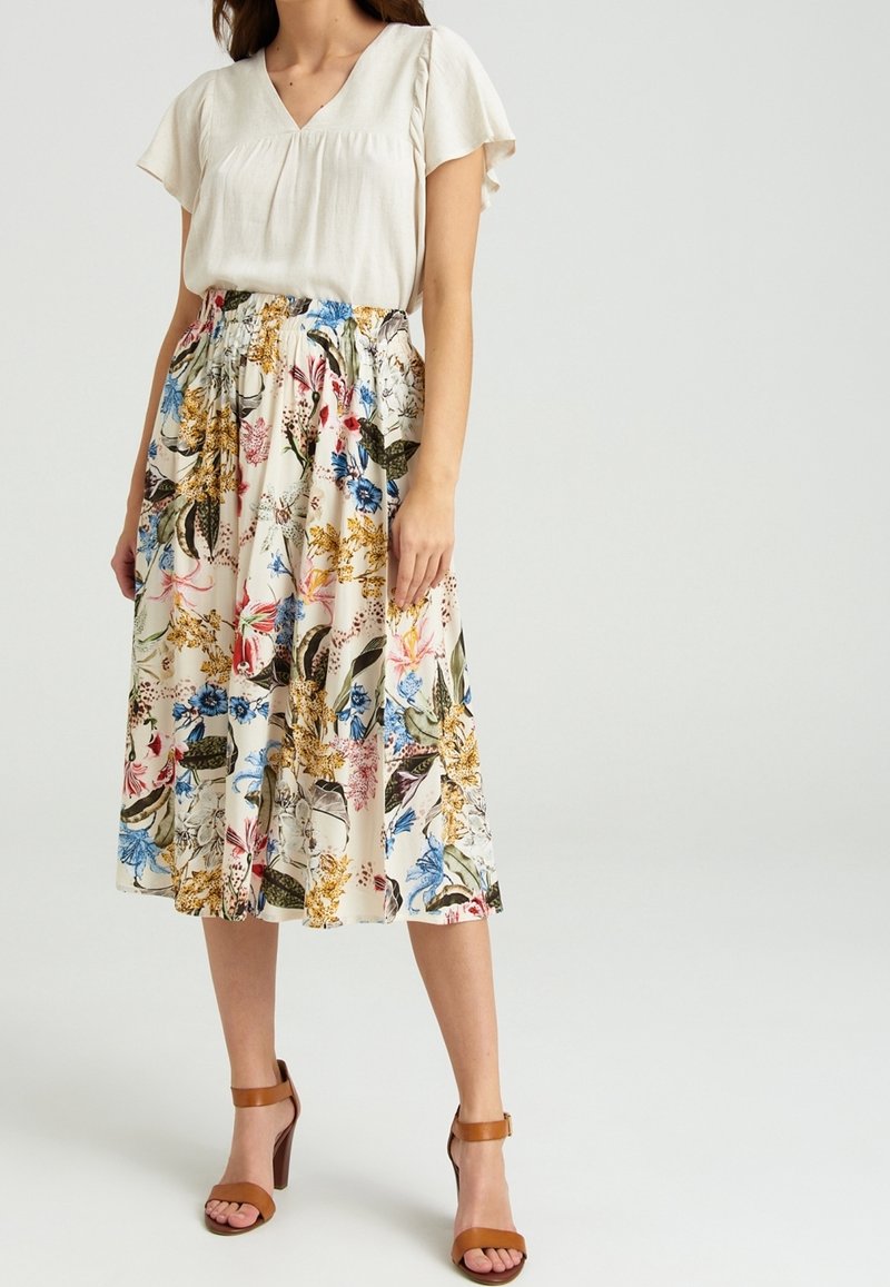 Lichte, beige blouse met korte, gerimpelde mouwen, gecombineerd met een bloemenrok tot de knie met blauwe, roze en gele patronen. Bruine, hoge sandalen.