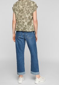 Koszulka z krótkim rękawem w kwiatowy wzór w oliwkowym odcieniu, zestawiona z jasnoniebieskimi dżinsami z prostymi nogawkami i podwiniętymi końcami oraz białymi sneakersami.