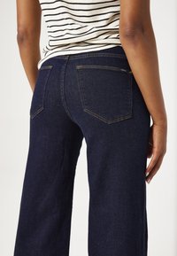 Jeans de pierna ancha en azul marino, fabricados con denim. Incluyen bolsillos traseros con detalles de costura y una textura suave.