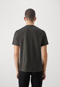 Homme aux cheveux courts et foncés portant un t-shirt uni gris foncé à manches courtes et un pantalon noir, debout face à un fond clair uni.