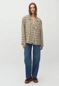 Beige kariertes Blazer mit doppelter Knopfleiste, breiten Revers und zwei Vordertaschen, kombiniert mit blauen weiten Jeans und dunklen Schuhen.
