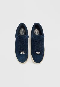 Σκούρα μπλε σουέντ αθλητικά παπούτσια με επίπεδη σόλα, στρογγυλή μύτη και κορδόνια. Διαθέτουν μεταλλικό λογότυπο στο μπροστινό μέρος και υφή στην επιφάνεια.