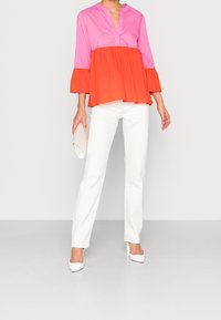 Top a blocchi di colore con parte superiore rosa, parte inferiore arancione e maniche a campana; abbinato a jeans bianchi e tacchi bianchi; tiene una pochette color crema.