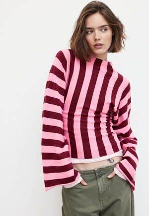 Jeune femme aux cheveux courts et bruns portant un haut rayé rose et rouge foncé à manches longues et un pantalon vert olive, les mains dans les poches.