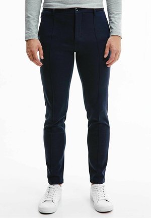 Hombre vistiendo pantalones ajustados azul marino, camisa de manga larga gris y zapatillas blancas, de pie con las manos relajadas a los lados.