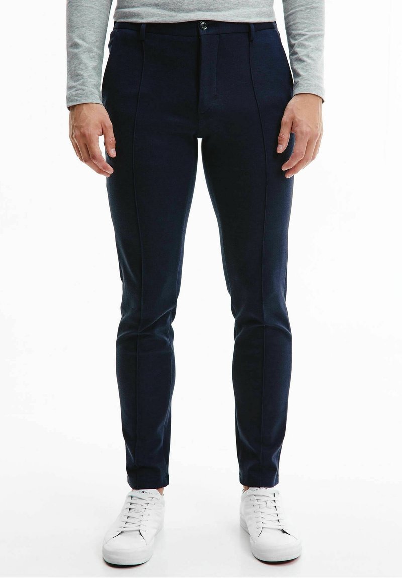 Homme portant un pantalon bleu marine ajusté, une chemise grise à manches longues et des baskets blanches, debout les mains détendues le long du corps.