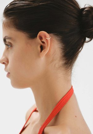 Pendiente de aro dorado en una oreja, con un diseño minimalista. El fondo es neutro, lo que resalta los detalles del pendiente.