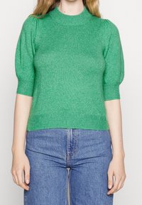 Groene gebreide trui met een hoge hals en korte pofmouwen, met een geribde zoom en een textiele stof. Gestyled met blauwe jeans.