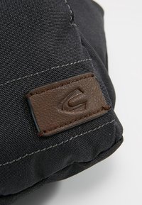 Coin d'un sac en tissu noir avec des coutures visibles et un patch en cuir marron cousu présentant un logo en relief incurvé.