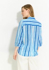 Camisa de rayas azul con un cuello tipo polo, tela semitranslúcida, mangas tres cuartos y un ajuste relajado. Rayas horizontales y verticales.