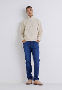 Sudadera de forro polar beige con media cremallera y un bolsillo frontal, combinada con vaqueros azules y zapatillas beige con acentos naranjas.