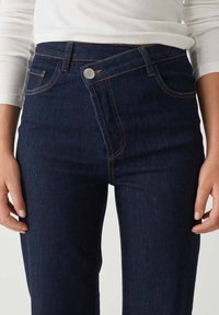 Close-up of dark blue denim jeans with an asymmetrical buttoned waistband worn with a white long-sleeve top.