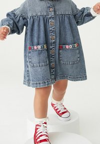 Jeanskleid mit Knopfleiste, Puffärmeln und zwei Taschen, verziert mit bunten floralen Akzenten. Kombiniert mit roten Hochschnitt-Sneakern.