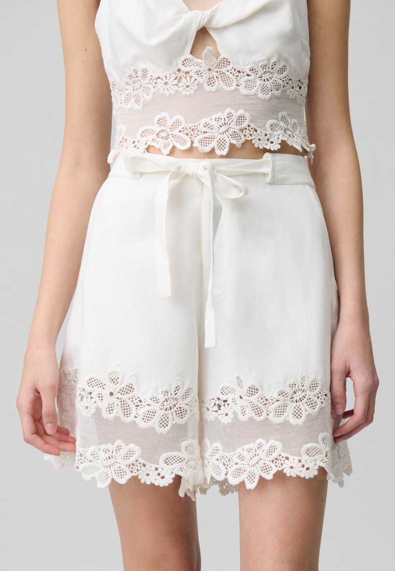 Set di top corto bianco e pantaloni a shorts, con bordi in pizzo e motivi floreali. Tessuto leggero con vita regolabile e dettagli a fiocco.