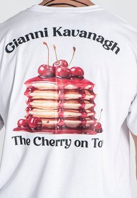 Camiseta de algodón blanca con un gráfico de apiladas de pancakes con cerezas y jarabe, el texto dice "Gianni Kavanagh" y "La Cereza en la Cima".