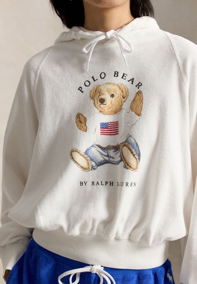 Hanorac alb cu un grafic al unui ursuleț de pluș care poartă un pulover cu steagul american, blugi albaștri și textul "Polo Bear de Ralph Lauren".