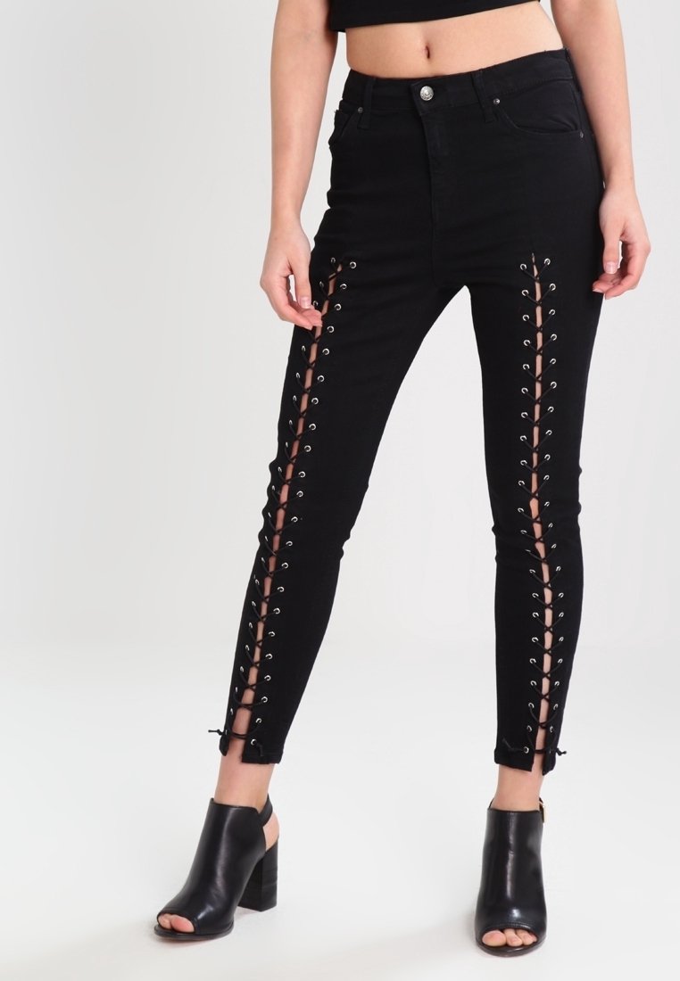 Femme portant un jean skinny noir taille haute avec des côtés lacés de la taille à la cheville, associé à des chaussures noires à talons blocs ouvertes à l'avant.