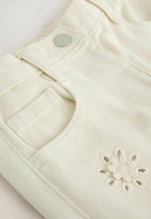 Tessuto di denim color crema con finitura strutturata, caratterizzato da un piccolo dettaglio ricamato a forma di fiore, bottone in metallo e design tradizionale delle tasche.