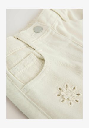 Kremasti traper tkanina s teksturiranim završetkom, s malim izvezenim cvjetnim detaljem, metalnim gumbom i tradicionalnim dizajnom džepa.