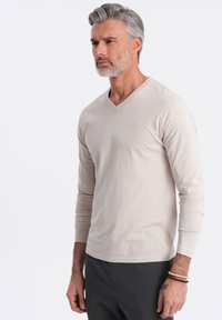 Beige Langarmshirt mit V-Ausschnitt aus weichem Stoff, mit lockerem Schnitt und glatter Textur, kombiniert mit dunklen Hosen.