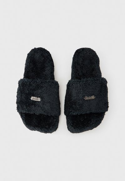 Juodos pūkuotos atviros basutės su plačia diržele ir maža metaline detale ant kiekvienos dirželės, išdėstytos ant balto fono.