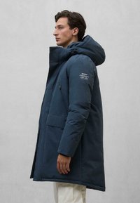 Granatowa kurtka puchowa z kapturem, zamkiem z przodu i kieszeniami po bokach. Materiał wydaje się gładki o matowym wykończeniu z delikatnymi detalami brandingowymi.